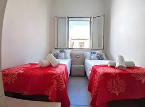 Habitación de hotel con dos camas, cada una con una colcha roja y almohadas blancas, una ventana con vistas y un espejo sobre las camas.