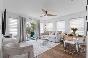 Ein modernes Wohnzimmer mit Sofa, Couchtisch, Esstisch und Stühlen. Es gibt einen Deckenventilator, einen flachbildfernseher an der Wand und ein Fenster mit Jalousien.