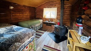 A cozy cabin with a bed, a small table, a stove, and a window. - Historical Cozy Off-Grid Cabin with Creekside Sauna near Log Cabin Creek, Tok Alaska (Tok)