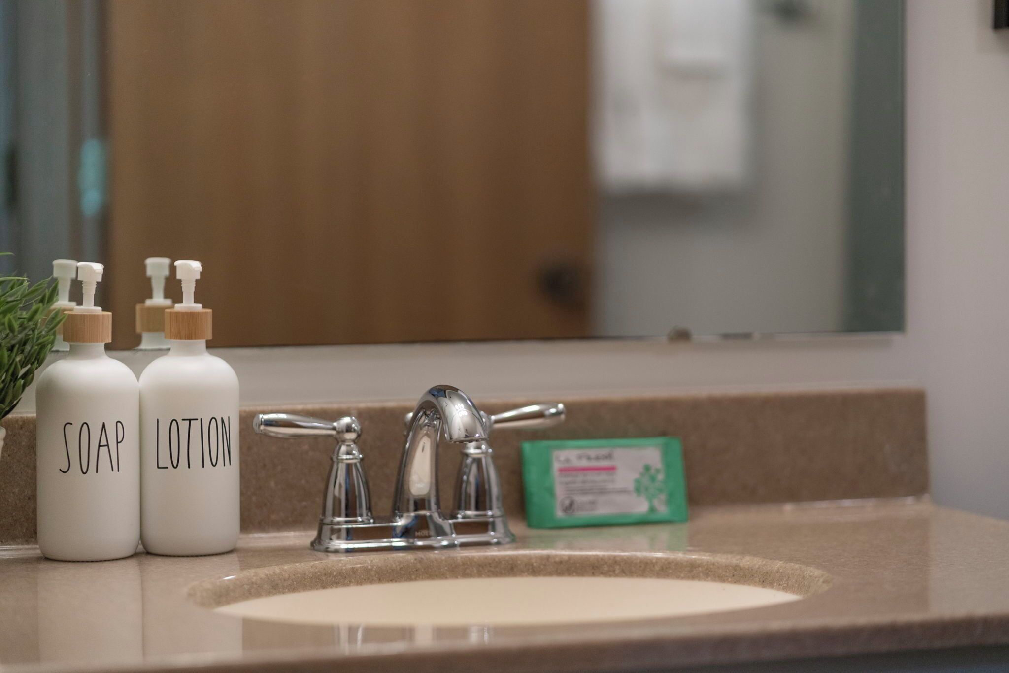 A bathroom sink with two bottles labeled 'SOAP' and 'LOTION' and a green card on the countertop.