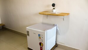 Un petit réfrigérateur blanc, une petite plante en pot posée sur une étagère en bois au-dessus, et un mur blanc en arrière-plan.