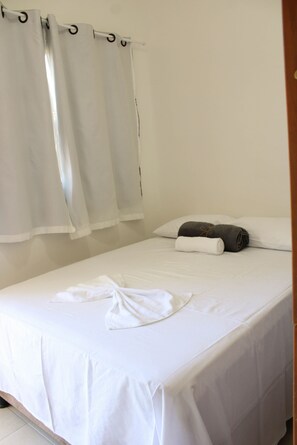 Una cama individual con ropa blanca, una toalla enrollada y un almohadón.