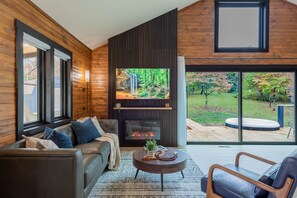 A modern living room with a fireplace, a sofa, a coffee table, and a view of a garden. - Aura Retreat | Hocking Hills, Romantic, Hot Tub (Creola)