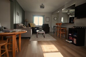 Ein Wohnzimmer mit Sofa, Couchtisch und Sesseln, ein Esstisch mit Stühlen, ein Bücherregal, ein Fernseher und eine Küchenzeile mit Kaffeemaschine.