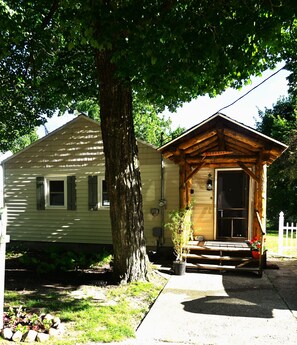 Ein Bungalow mit Veranda, ein großer Baum davor und eine kleine Gartenhütte.