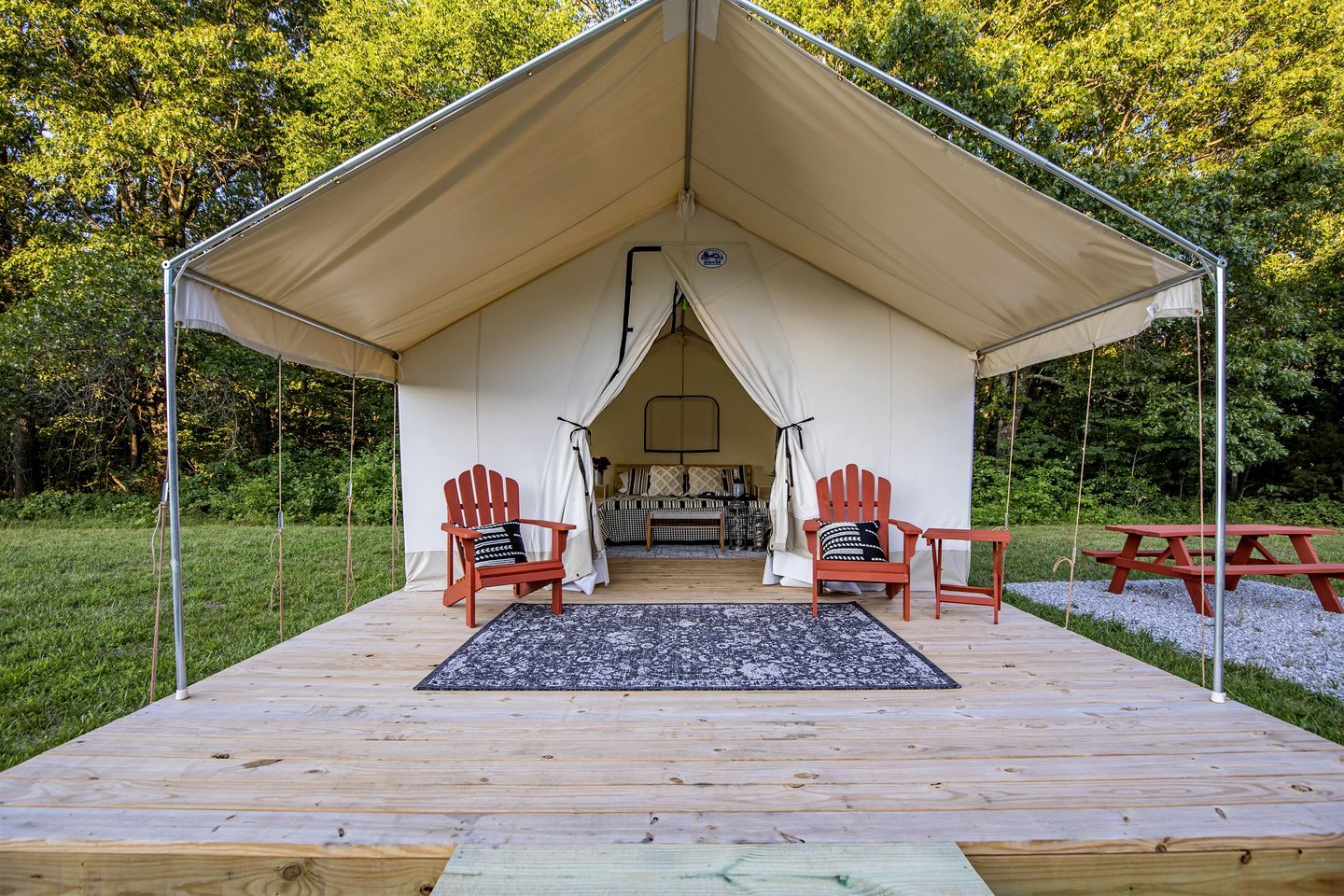 A tented accommodation with a wooden deck, two red Adirondack chairs, a picnic table, and a bed with pillows.