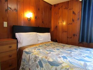 A bedroom with wooden walls, a bed with a floral bedspread, a nightstand, and a wall-mounted light.