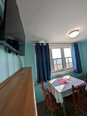 A room with a wooden table, a television mounted on the wall, a window with curtains, and a view of the sea.
