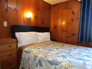 Ein Schlafzimmer mit Holzwänden, einem Bett mit floralem Überwurf, einem Nachttisch und einer Wandleuchte.