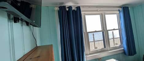 A room with a wooden table, a television mounted on the wall, a window with curtains, and a view of the sea.