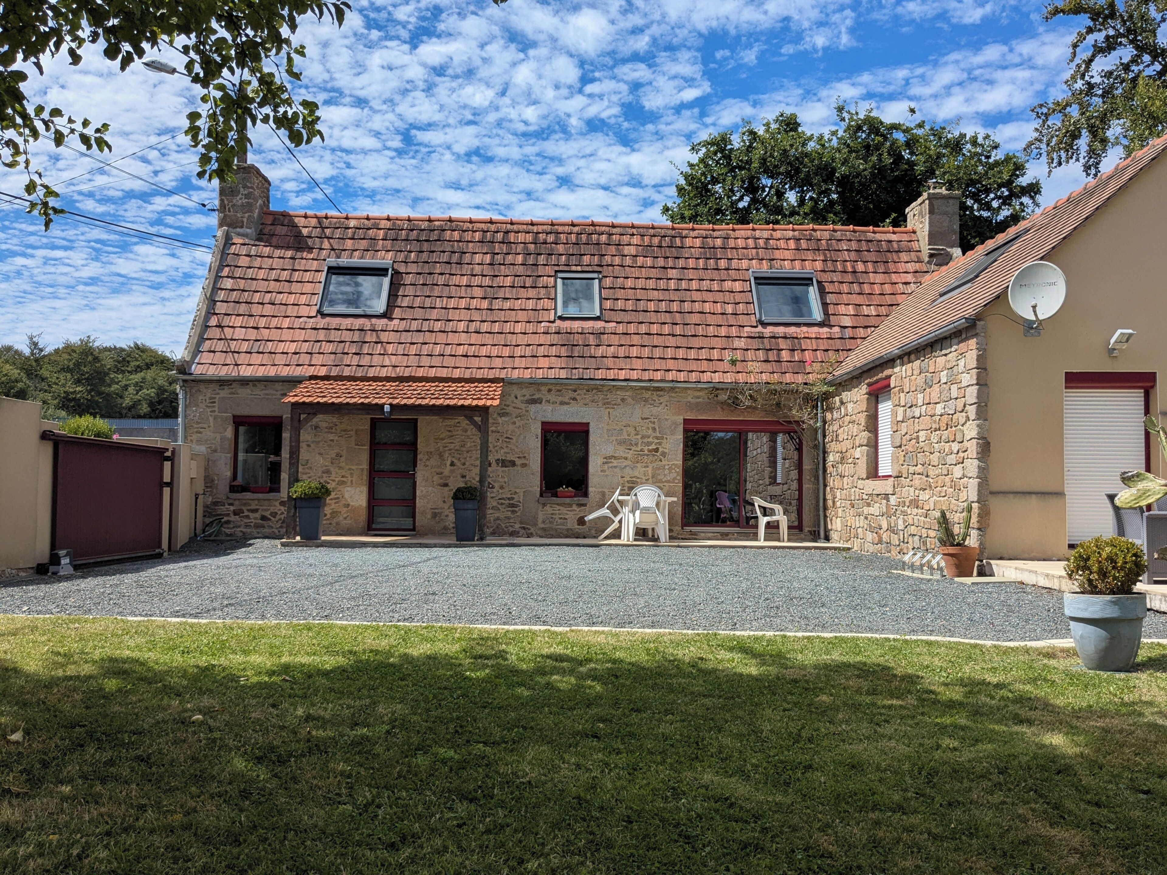 A stone house with a tiled roof, a gravel driveway, and a small patio with chairs.
