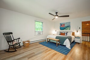 Ein Schlafzimmer mit einem Bett, einem Schaukelstuhl, einem Deckenventilator, einem Fenster mit Jalousien, einem Gemälde und einem Nachttisch mit Lampe.