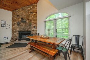 A dining area with a stone fireplace, wooden table, benches, and a view through a window with blinds. - Quaint Retreat w/ Fire Pit & Hot Tub! (McHenry)