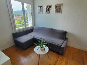 Ein Eckzimmer mit grauem Ecksofa, einem kleinen weißen Tisch mit einer Pflanze und zwei gerahmten Kunstwerken an der Wand.