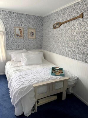 A neatly made bed with white bedding, a wooden headboard, and a small bedside table with a book. The room has patterned wallpaper and a window with curtains. - Cozy Lakefront Cabin with Pontoon and Paddleboards Near Fremont, Indiana (Fremont)
