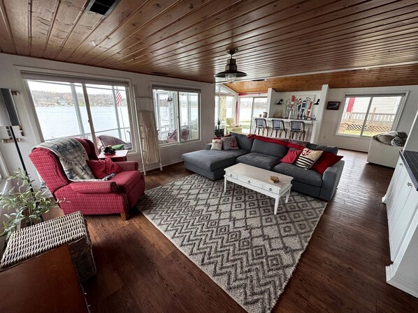 Una sala de estar acogedora con sofá, sillón y mesa de centro, ventanas amplias con vistas a un lago, y suelo de madera.