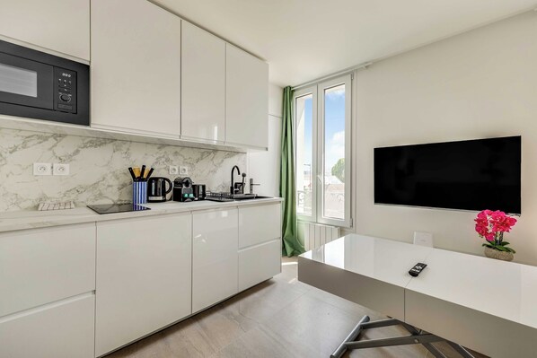 Una cocina moderna con armarios blancos, microondas integrado y cafetera. Hay un ventanal con cortinas verdes, un televisor de pantalla plana y una pequeña mesa con un florero.