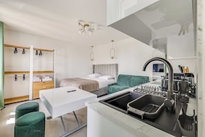 Ein kleines Hotelzimmer mit einer Küchenzeile, einem Bett mit Kopfteil, einem kleinen Tisch und einem grünen Hocker.