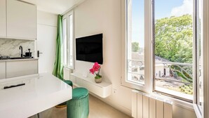 Una cocina moderna con encimera blanca, una silla verde, un televisor montado en la pared y una ventana con vistas a zonas verdes y edificios.