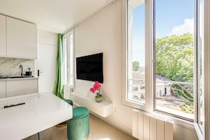 Una cocina moderna con encimera blanca, una silla verde, un televisor montado en la pared y una ventana con vistas a zonas verdes y edificios.