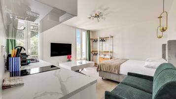 A modern hotel room with a kitchen area, a bed, a sofa, and a TV.