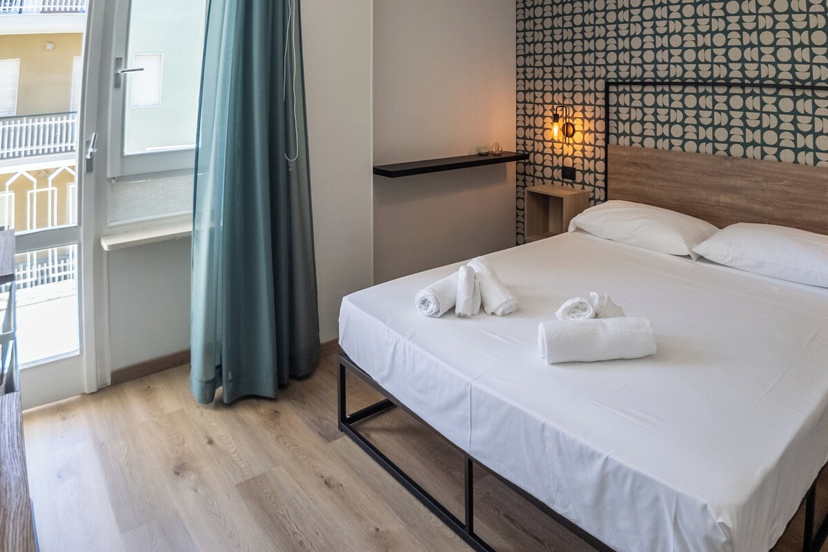 Habitación de hotel con cama, ventana con cortinas, pared con diseño circular y mesita de noche con lámpara.
