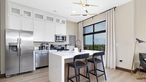 Cocina moderna con armarios blancos, electrodomésticos de acero inoxidable, una isla grande con taburetes y vistas a través de un ventanal.