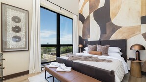 Una habitación de hotel moderna con una cama grande, un banco, una mesita de noche con una lámpara, un ventanal con vistas y una obra de arte en la pared.