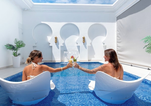 Deux personnes dans un bateau blanc, dans une piscine avec un mur blanc et une plante.
