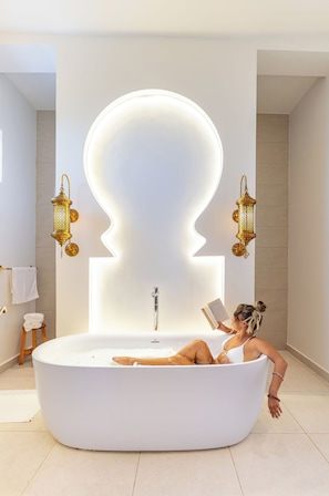 Une femme se détend dans une grande baignoire blanche, agrémentée d’un décor mural original, avec un livre à portée de main et deux appliques murales décoratives.