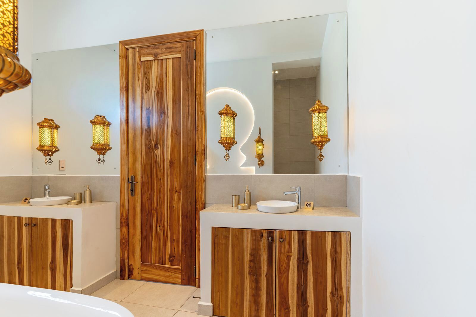 Un baño con dos lavamanos, armarios de madera, una puerta y faroles en la pared.