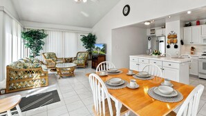 Un vaste espace de vie comprenant un coin repas avec une table pour quatre personnes, entouré de meubles en osier, d’un ventilateur de plafond et d’une grande fenêtre équipée de stores.