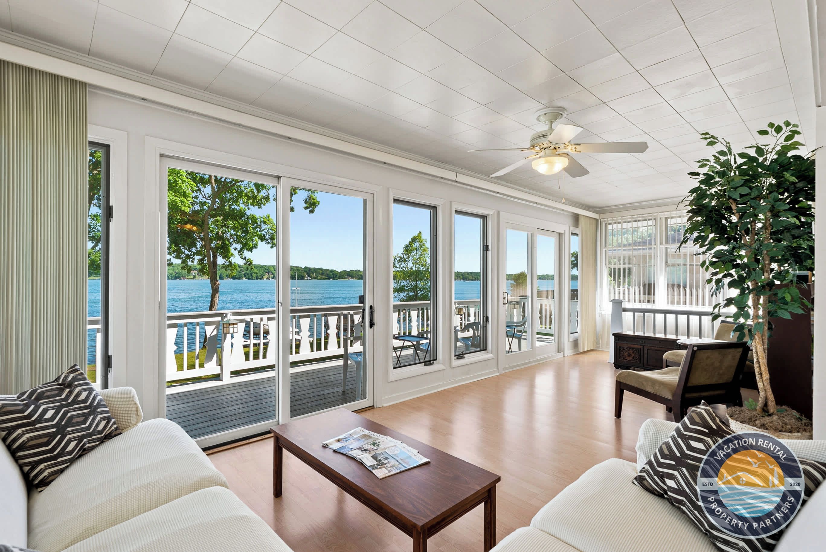 A spacious room with a large sliding glass door leading to a balcony with a view of the water. There is a ceiling fan, a wooden table, and a sofa with patterned pillows.
