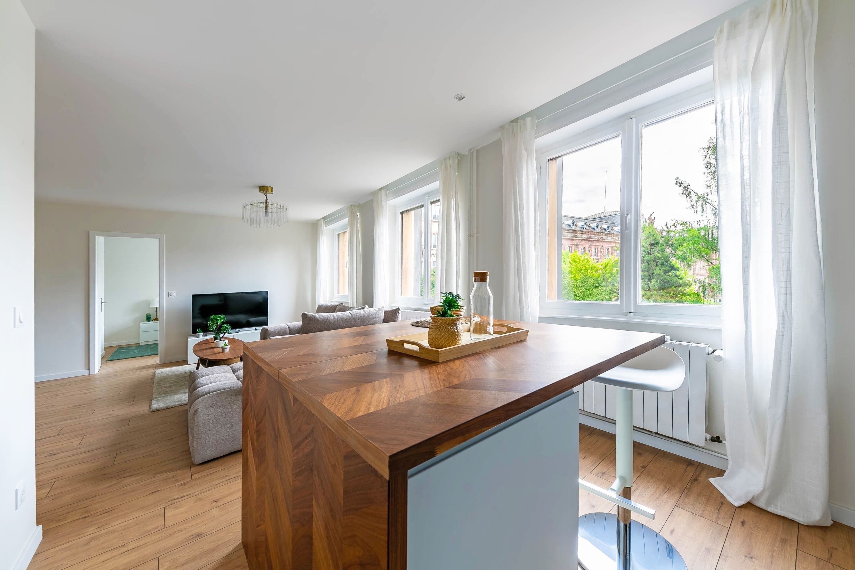 Un salon moderne avec une table à manger en bois, un canapé, un téléviseur et de grandes fenêtres dotées de rideaux.