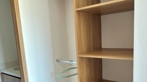 A wooden wardrobe with three shelves and a hanging rod, located in a room with white walls and tiled flooring.