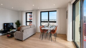 Una sala de estar moderna con sofá, mesa de comedor, sillas, televisor y ventanas amplias con vista a la ciudad.