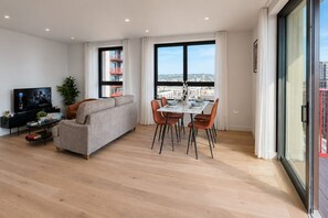 A modern living room with a sofa, dining table, and chairs, a TV, and large windows with a city view. - Modern Greenwich flat with Balcony near The O2 & Canary Wharf (London)