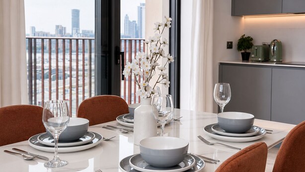 Mesa de comedor dispuesta con platos, tazones, copas de vino y cubiertos, con un jarrón de flores y vista a la ciudad a través de amplias ventanas.