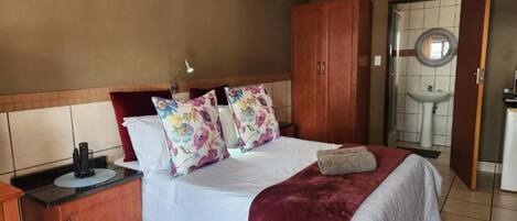 A bedroom with a bed, a wooden wardrobe, a bedside table, and a bathroom with a sink and mirror.