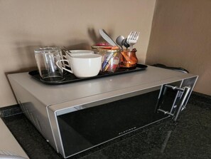 Una zona de cocina con un microondas, una bandeja con tazas, un tarro y utensilios.