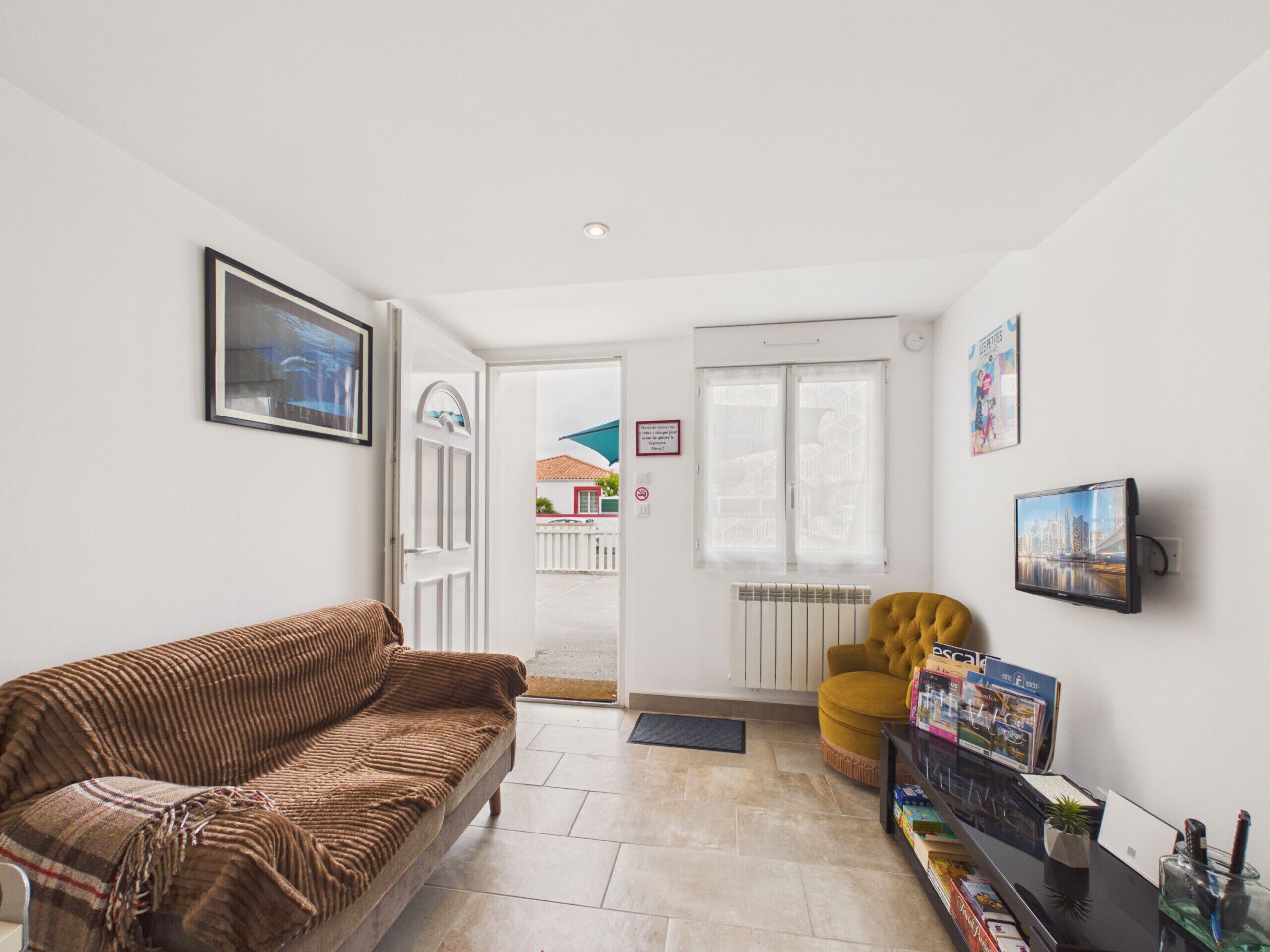 Een woonkamer met een bruine bank, een gele fauteuil, een aan de muur bevestigde televisie, en een deur naar buiten.