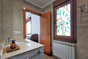 Um banheiro com porta de madeira, janela com padrão de folha, pia com dosador de sabão e radiador.
