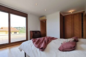 Um quarto com uma janela ampla, uma cama com almofadas, um guarda-roupa de madeira e uma pequena mesa.