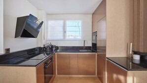 Cuina compacta amb taulell negre, armaris de fusta, forn de pared i finestra amb persianes.