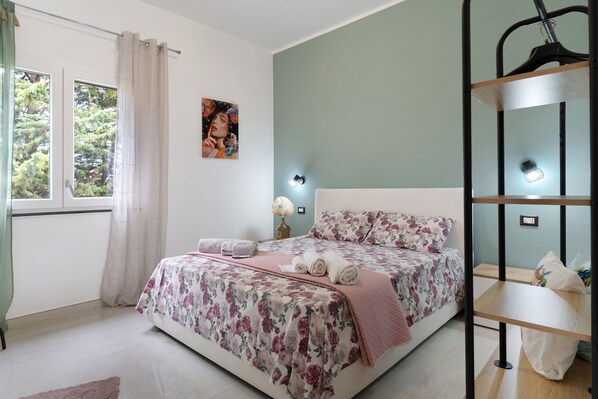 Un dormitorio con cama, ventana con cortinas, estante en la pared y un cuadro.