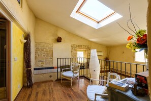A room with a sloped ceiling, a skylight, a stone wall, a wooden floor, a white chair, a white table, and a book titled 'MAXIME CITTAM'. - Les Clés des Garrigues - Maison climatisée (Fournes)