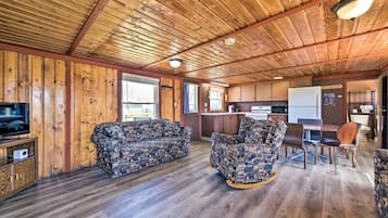 A cabin interior with a wooden ceiling, a sofa, a patterned armchair, a dining table with chairs, a kitchen area, and a television on a wooden cabinet.