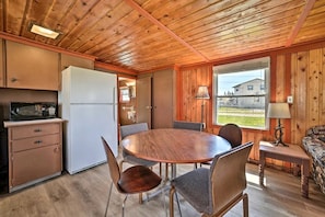 Interior de una cabaña con techo de madera, una mesa redonda para comer, sillas, un refrigerador, un microondas y una ventana con vista a una casa.