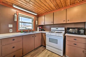 A kitchen with wooden cabinets, a stove, a microwave, and a window with a view. - Waterfront Cabin with Kayaks, Dock & Fire Pit in Houghton Lake, Michigan (Houghton Lake)
