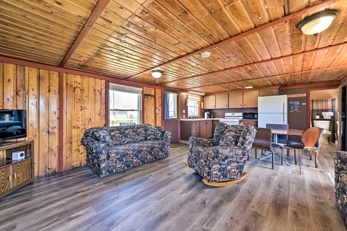 A cabin interior with a wooden ceiling, a sofa, a patterned armchair, a dining table with chairs, a kitchen area, and a television on a wooden cabinet.